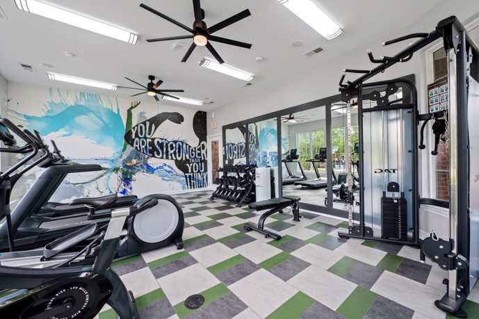 Community fitness center with white walls and ceiling fans, grey, white, and green tiled carpeting and treadmills and stationary bikes facing mirrors
