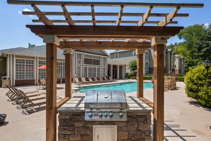 Outdoor stone gas grill of an apartment grilling station  under a wooden pergola and overlooking the swimming pool and deck chairs