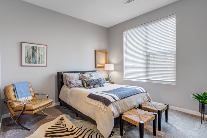 An inviting apartment bedroom adorned with plush carpeting, featuring a comfortable queen bed, two stylish stools, a leather chair for relaxation, and a window allowing in natural light, all within Views of Music City.