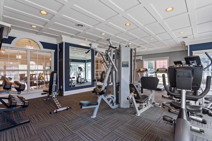 A carpeted fitness center within Spring Hill, equipped with a variety of gym equipment, enhanced by large windows allowing abundant natural light, and mirrors to facilitate workouts.