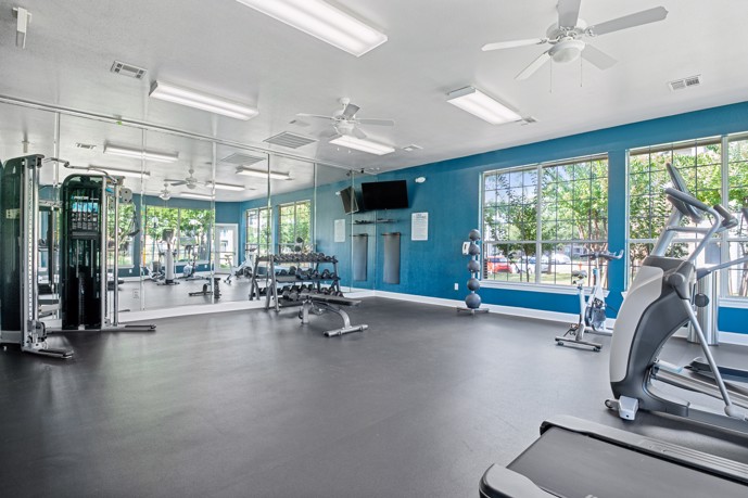 Well-lit fitness center boasting expansive windows, state-of-the-art cardio and weight equipment, mirrors for proper form, and refreshing ceiling fans.