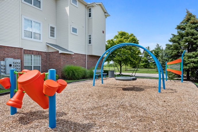 Outdoor community playground on a wood chip ground with apartment buildings to the left and trees in the distance