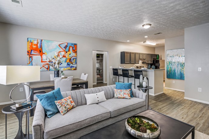 A modern open floor plan apartment with a two-seater grey couch, a dining area, and a kitchen with a bar counter at The Commons at Canal Winchester. 