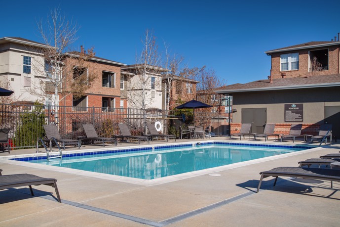 Outdoor swimming pool surrounded by deck chairs with a pool house on the left and a fence in the back with apartments in the background