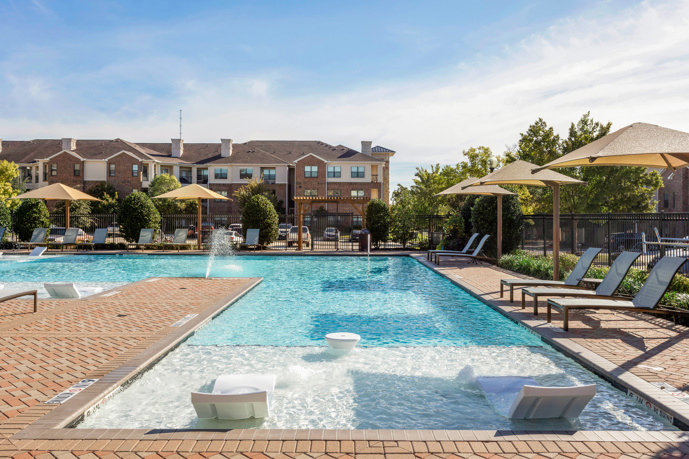 An outdoor community swimming pool in a brick patio with deck chairs and the apartment buildings around it