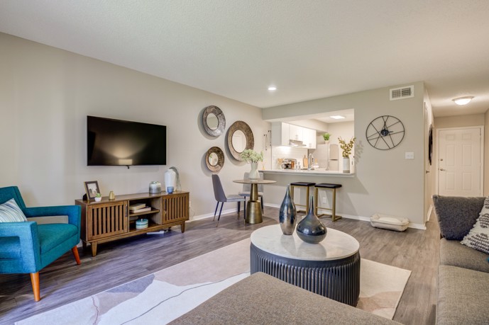 An inviting open-floor plan living space at The Invitational, featuring comfortable furniture in the living room, a cozy dining area with a quaint table, and a glimpse into the well-appointed kitchen through a window.