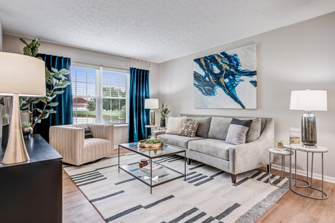 Grey furnished apartment with hardwood floors, an area rug, seating, coffee table, table lamps, and blue drapes, and abstract artwork on the wall