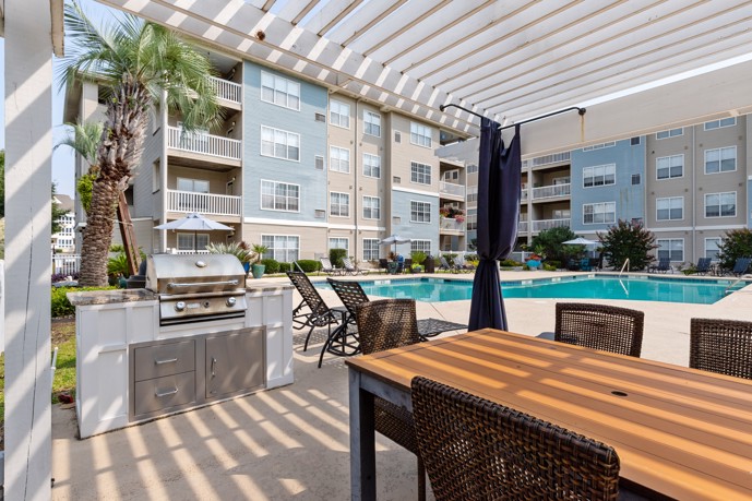 View of the swimming pool at Cherry Grove Commons apartments in North Myrtle Beach, SC from the outdoor grilling station and dining area under a white pergola
