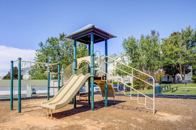 Playground adorned with monkey bars and slides, adjacent to verdant grass, a picnic bench, grill, and charming apartment buildings, providing recreation for residents.