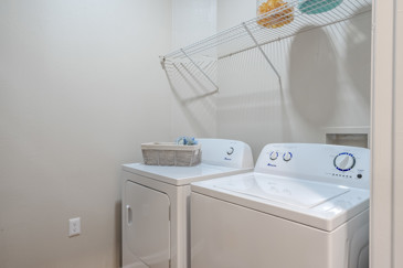 An apartment laundry room at The Enclave at Tranquility Lake in Riverview, FL, equipped with a washer and dryer, shelving, and bright overhead lighting.