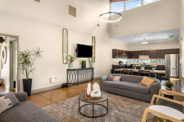 Long view of an apartment clubhouse with a large seating area by fireplace in the foreground, kitchen area in the background, and an exposed wooden beam high ceiling 