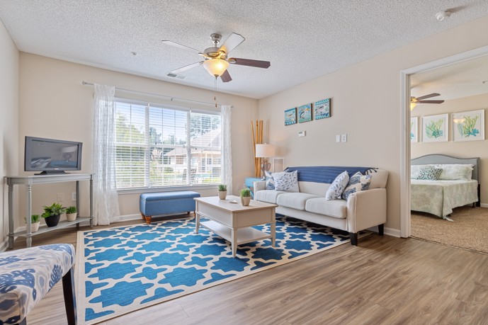 Light tan living room with wood-style flooring, a blue area rug, seating area, an open door to a bedroom at Cherry Grove Commons apartments in Sunset Beach, SC