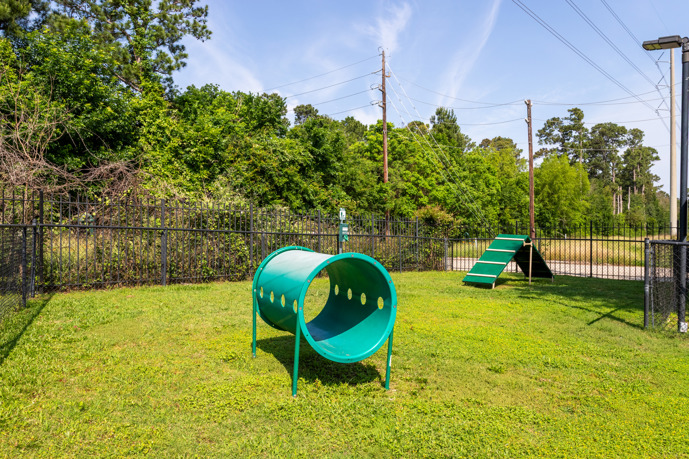 Fenced-in grass apartment community dog park surrounded by an iron fence and trees, featuring obstacles for the dogs