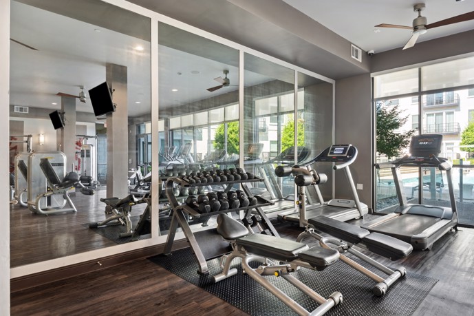 A state-of-the-art fitness center at VV&M apartments, featuring gym equipment, mirrors, large windows, and wood-style flooring, providing residents with a stylish and comfortable space to stay active and healthy.