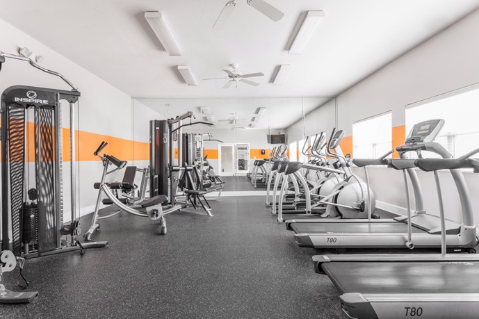 Fitness center equipped with cardio and weight machines, boasting ample natural light from large windows and enhanced with cooling ceiling fans.