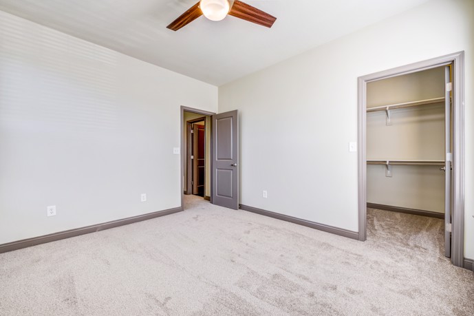Empty apartment bedroom with grey walls and carpeting, a ceiling fan, closet, and door opening to another room