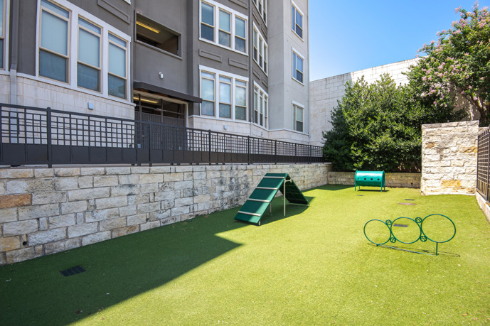 A fenced dog park located in front of the VV&M apartments in Dallas, Texas, featuring artificial grass and dog equipment, providing a safe and fun space for residents' furry friends to play and exercise.