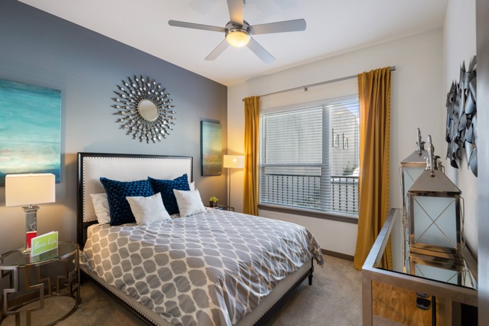 A comfortable bedroom at VV&M apartments in Dallas, Texas, featuring a carpeted floor, a queen-sized bed, two side tables, a window, and an armoire, providing residents with a relaxing space to rest and recharge.