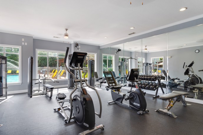 Community fitness room with exercise equipment and dumbbells with  a mirrored wall to the right and windows and a door overlooking the outdoor pool ahead