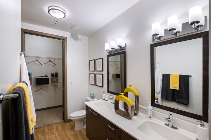 A modern bathroom at VV&M apartments, featuring dual sinks, wood-style flooring, large mirrors, and a separate closet.
