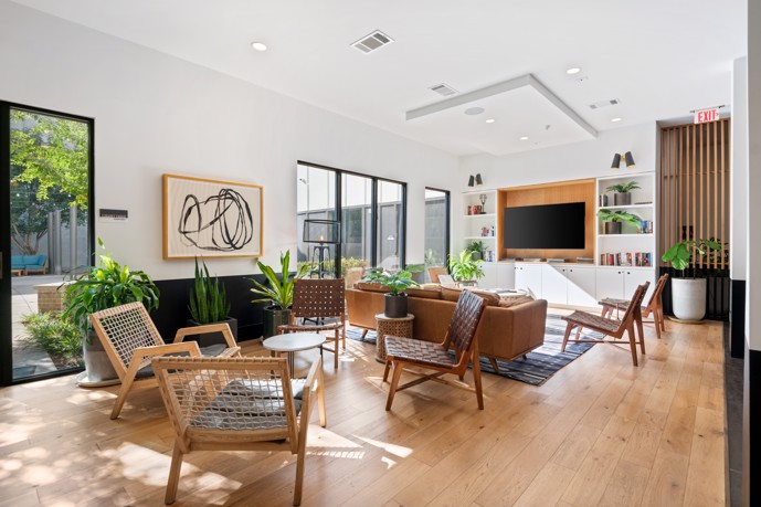 An inviting community lounge, boasting ample natural light through its large windows, two distinct seating areas, and a centrally placed TV for relaxation and entertainment.