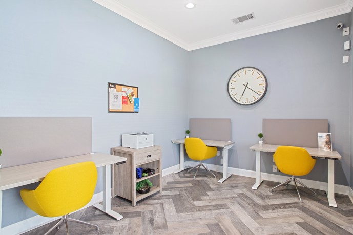 Community work stations with three yellow chairs at grey desks and a large round clock on the wall