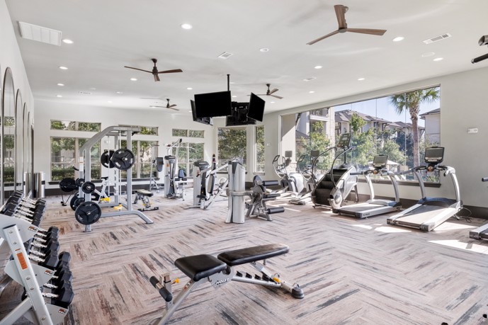 Well-equipped fitness center, featuring multiple ceiling fans, TVs, expansive windows, and an array of exercise equipment for residents' wellness and enjoyment.
