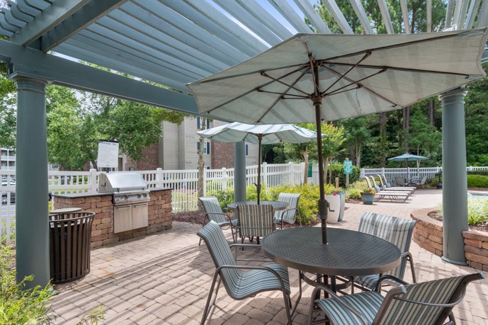 Outdoor community poolside picnic area equipped with tables, chairs, umbrellas, and a barbecue grill nestled under a pergola structure.