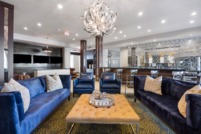 The luxurious clubhouse at VV&M apartments, featuring lavish blue couches, a kitchen bar, and elegant chandeliers, providing residents with a comfortable and stylish space to relax and socialize.