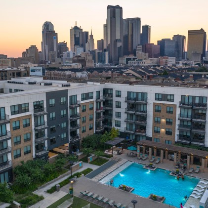 A captivating aerial perspective showcasing Eleven10 at Farmers Market, highlighting the apartment building, the sparkling pool area, and the iconic downtown Dallas skyline.