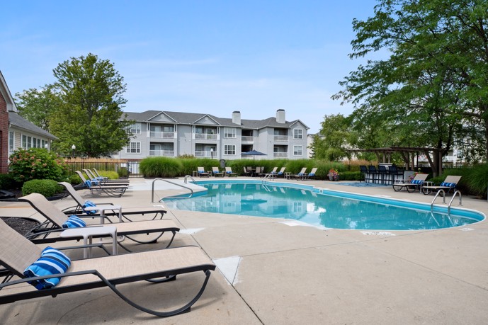 Outdoor apartment community swimming pool with deck chairs to the left, a large tree to the right and apartment buildings in the distance