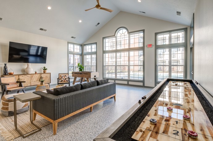 Apartment clubhouse with vaulted ceilings, a seating area facing a flat screen tv, large windows along one wall, and a table shuffleboard game