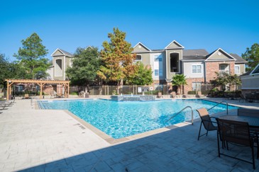 Large grey paved patio and swimming pool with apartment building behind it and a patio table with chairs in the shade in the foreground