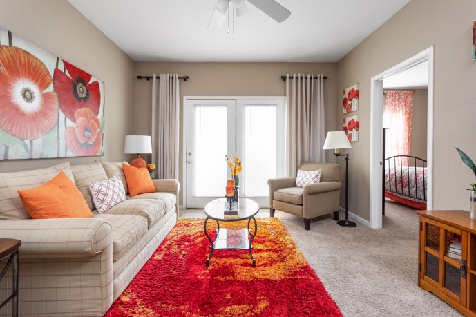 A tan apartment living room with comfortable seating, beige wall-to-wall carpeting, a coffee table, lamps, and a red area rug and matching wall art