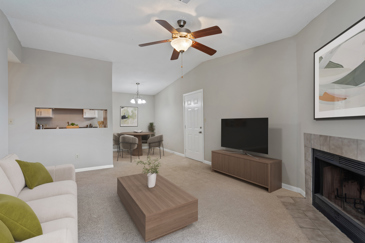 A modern carpeted living room with a fireplace and a kitchen next to it at the Villages at Spring Hill. 