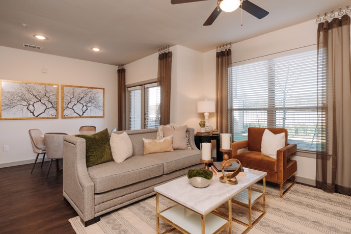 Inviting living room boasting ample natural light from large windows, complemented by a refreshing ceiling fan, adjacent to a cozy dining area featuring hardwood-style flooring.
