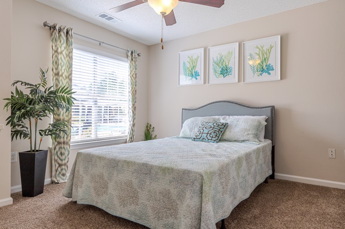Tan bedroom with a window, large plant, and floral artwork over the bed at Cherry Grove Commons apartments in North Myrtle Beach, SC