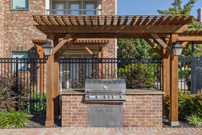 Outdoor brick gas grill under a pergola in front of a black iron fence and an apartment building behind it 