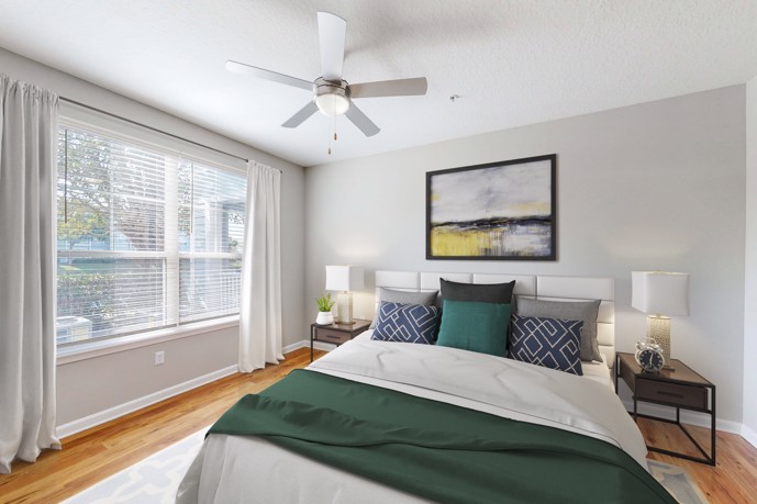 A cozy bedroom with wood flooring, featuring a queen bed flanked by two side tables, bathed in natural light from large windows, offering residents a serene retreat within The Enclave at Tranquility Lake.