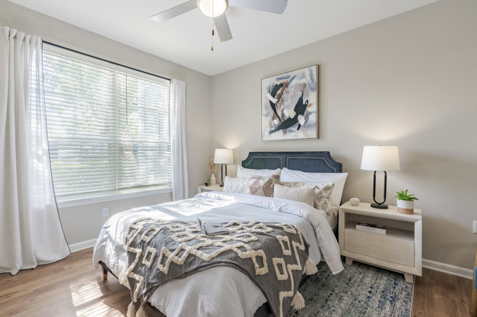 A cozy bedroom with wood flooring, featuring a queen bed flanked by two side tables, bathed in natural light from large windows, offering residents a serene retreat within The Enclave at Tranquility Lake.