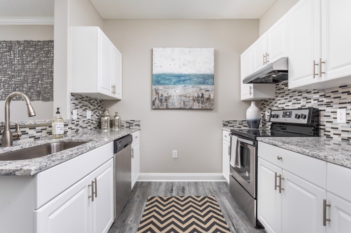 A modern galley kitchen at The Village at Auburn, featuring white cabinets, wood-style flooring, elegant granite countertops, and a stylish backsplash, providing residents with a functional and stylish space to cook and entertain guests.