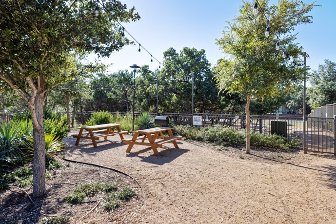 Outdoor area adorned with verdant plants, two inviting picnic tables, charming string lights, and a convenient barbecue grill.