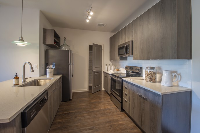 A modern apartment kitchen with grey cabinets, quartz countertops, stainless steel appliances, and wooden floors