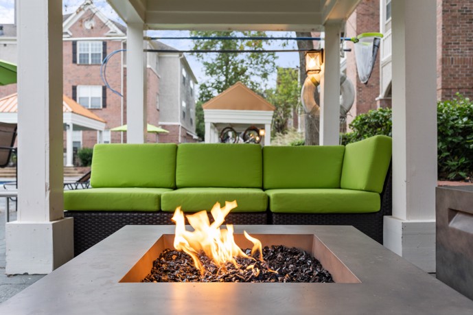 Outdoor fireside lounge with a lit fire pit in the foreground and a green sofa facing it under a pergola and apartments in the background