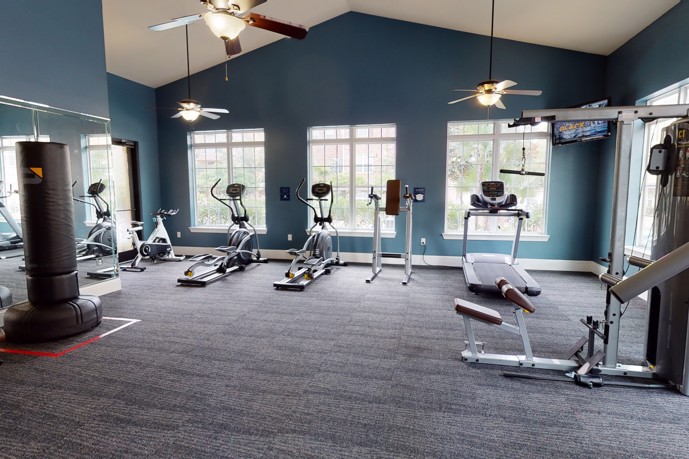 A well-equipped gym featuring cardio and weight equipment, large windows for natural light, and ceiling fans for comfort.