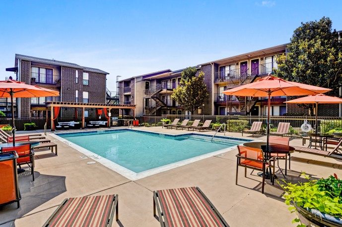The Vue at Knoll Trail apartments have an expansive pool area surrounded by lush greenery, complete with comfortable loungers, umbrellas, and lounge seating under a pergola, creating a serene and inviting space.