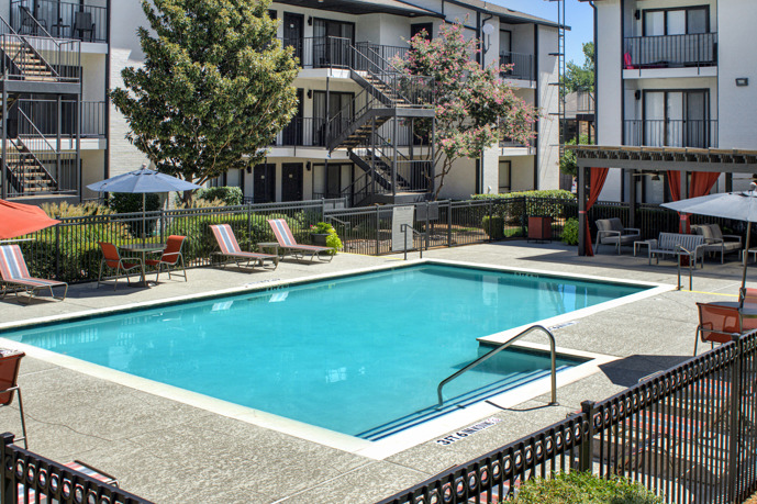 The Vue at Knoll Trail apartments have an expansive pool area surrounded by lush greenery, complete with comfortable loungers, umbrellas, and lounge seating under a pergola, creating a serene and inviting space.