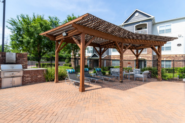 Outdoor wooden veranda with chairs under it and sun shining through it to create shadows and a grilling station behind it to the left