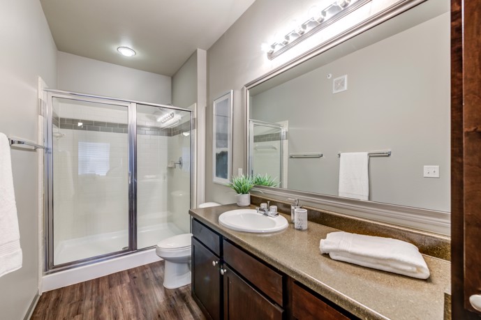An apartment bathroom with light-colored walls, a dark wooden vanity, large mirror, and glass door shower