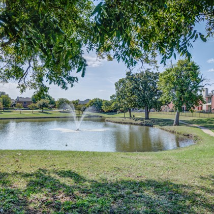 A picturesque pond featuring a tranquil fountain, encircled by a winding walking pathway and lush trees at Lakeside at Coppell.